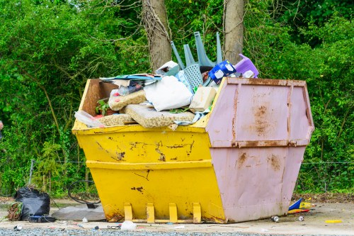An overflowing large skip bin situated outdoors on a gravel surface, with a vibrant yellow base and a contrasting pink lid. The bin is filled with a mixture of household waste including plastic containers, a white plastic bag, foam packaging, fabric textiles, and other miscellaneous rubbish. The contents spill slightly over the top edges, with some items leaning against the sides, indicating it is nearly full. The background features dense green foliage with trees and bushes, suggesting a suburban or industrial area near Woolwich. The lighting is natural, illuminating the various textures and colors of the waste materials clearly. Nearby, a small black plastic bag and some scattered debris can be seen on the ground, emphasizing the dump's presence. Business Waste Removal Woolwich provides rubbish removal services, evident from the environmental context and the type of waste stored in the skip bin, which is typical for managing waste removal needs within the local area. The scene reflects a typical outdoor rubbish collection point ready for disposal or collection services.