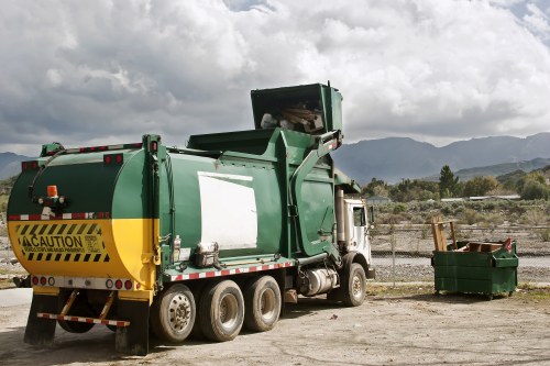 A large green and yellow rubbish collection truck, parked on an uneven dirt surface, is positioned with its rear compactor door open, indicating active waste loading. The truck is designed for waste management services, featuring a sturdy, metallic exterior with visible rivets and a caution sign near the rear. To the right of the truck, a green, partially open waste disposal bin is placed on the ground, filled with miscellaneous waste materials. The environment appears to be an outdoor waste disposal or transfer site, with a backdrop of rugged landscape, distant mountains, and a partly cloudy sky overhead. The scene reflects a professional rubbish removal operation, possibly within the Woolwich area or nearby in south-east London, aligning with services offered by Business Waste Removal Woolwich. The overall setting is utilitarian, focused on waste collection activities with natural lighting highlighting the vehicle and bin in the foreground, amidst a typical outdoor waste management environment.