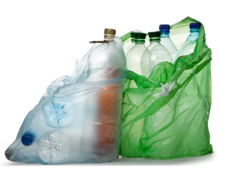 The image displays a collection of empty plastic bottles and a plastic bag, primarily consisting of transparent and semi-opaque plastic materials. The bottles vary in size and shape, with some featuring coloured caps in shades of blue and beige, and others without caps. The bottles appear to be made of PET plastic, exhibiting a glossy, smooth surface with visible creases and folds from handling. The bottles are positioned upright, some leaning against each other, with the majority grouped together outside of any container. A green plastic shopping bag, crumpled and open at the top, surrounds the bottles on the right side, adding a contrasting color and texture to the clear plastic objects. The overall lighting highlights the reflective qualities of the plastic, creating subtle glare and shadows. The background is plain white, emphasizing the objects themselves and their status as waste suitable for rubbish removal services, such as those offered by Business Waste Removal Woolwich, serving local recycling and waste management needs in Woolwich or nearby areas.