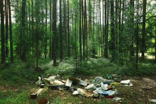A pile of domestic and recyclable waste, including cardboard boxes, plastic bags, and miscellaneous household rubbish, is collected on a dirt ground in a forested area with tall, slender trees and dense green foliage in the background. The rubbish appears to be discarded improperly, lying disorganized amidst natural surroundings, highlighting environmental concerns related to littering. The sunlight filters through the tree canopy, casting dappled light over the waste and the forest floor. This scene illustrates the importance of proper rubbish disposal practices and the role of waste management services, such as those provided by Business Waste Removal Woolwich, in maintaining clean and environmentally responsible communities within areas like Woolwich, London, and nearby postcode districts. The contrast between the natural environment and the scattered waste underscores the need for effective rubbish removal in rural and urban settings alike.