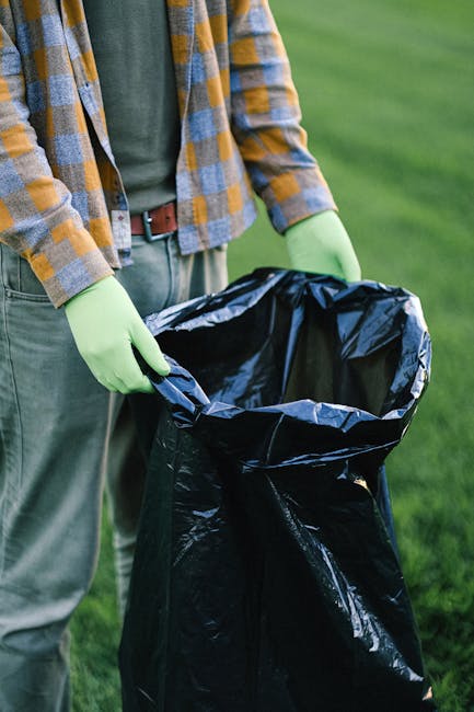 A person standing outdoors on a grassy area, wearing a checkered shirt in shades of yellow, blue, and gray, along with green gloves, is holding open a large black garbage bag made of shiny, crinkled p