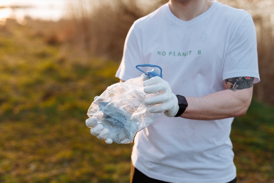 A person standing outdoors on a grassy area, wearing a checkered shirt in shades of yellow, blue, and gray, along with green gloves, is holding open a large black garbage bag made of shiny, crinkled p